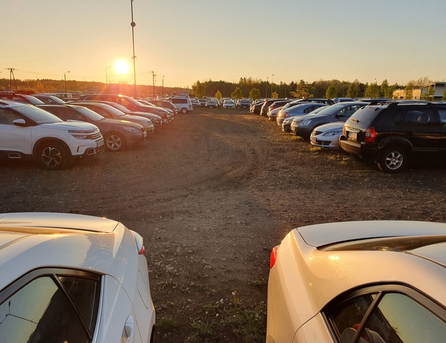 KTW Parking - Szeroki plac pełen zaparkowanych samochodów. Zachód słońca oświetla auta. Parking jest nieutwardzony.