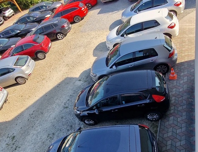 Parkovisko PARK4YOU - Plné áut parkovisko na kamenistej povrchu. Areál obklopený zeleňou.