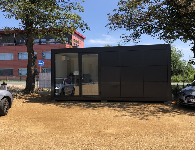 Parkovanie na dovolenke – parkovanie s kamienkovou plochou. Naľavo a napravo stoja autá. V pozadí sa nachádza kancelársky kontajner.