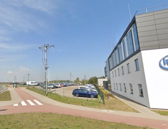 Parkplatz Piątka - Parkplatz mit Zaun neben dem Gebäude und Übergangsschild. Auf dem Parkplatz stehen geparkte Autos.