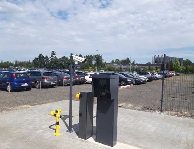 Parking LOT 24 – Wjazd z szlabanem i kamerą monitoringu. Samochody stoją w rzędach na utwardzonym placu. Parking jest ogrodzony.