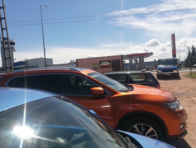 Parking Na Wylocie - kolorowe auta stoją na parkingu. Teren jest nieutwardzony, niezadaszony. W tle widać stację paliw.