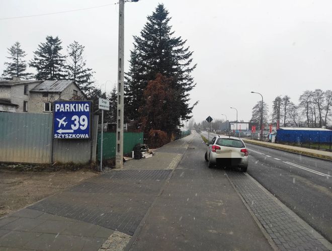 Parking Szyszkowa 39 - Po lewej widoczny jest znak parkingu. W tle stoją drzewa i budynek. Na drodze zaparkowany jest samochód.