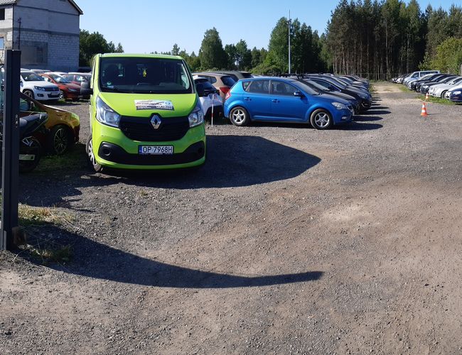 Gran Estacionamiento - El terreno está cercado y gravilloso. Se pueden ver coches estacionados y una furgoneta verde. Un cartel informa sobre el nombre y contacto del estacionamiento.