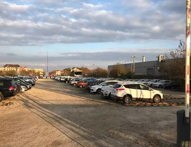 Żwirowy parking z dużą liczbą samochodów, szlabanem po prawej stronie i zabudowaniami w tle.