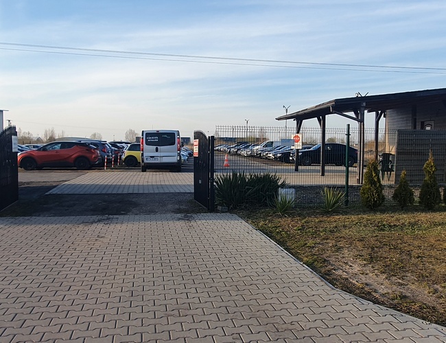 WIZ-PARK - El aparcamiento está cercado. La puerta está abierta. Se pueden ver coches aparcados y el edificio.