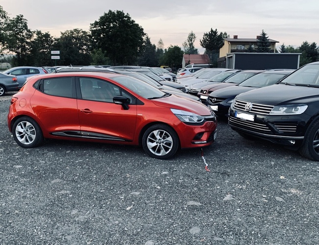 Krakow KING - Na štrkovom parkovisku je veľa áut. Obklopujú ho stromy a budovy. V popredí je červené auto.