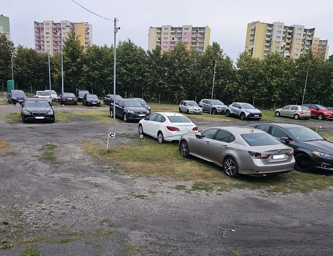 Odlotowy Parking BZG – Na zdjęciu widoczne są zaparkowane samochody. Teren jest otwarty i ma żwirową nawierzchnię. W tle są bloki mieszkalne.