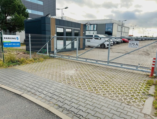 Parking Piątka – parking jest ogrodzony i utwardzony. W tle znajdują się biurowce.