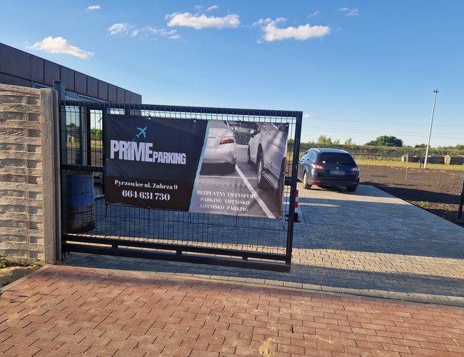 Parking Prime – Ogrodzony teren z bramą i tablicą informacyjną. Samochód wjeżdża na wybrukowany plac.