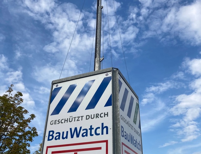 Parking P365 Valet - Wysoki maszt z kamerami monitoruje parking. Na dole widoczny jest znak BauWatch. Obok rośnie drzewo, a niebo jest niebieskie.