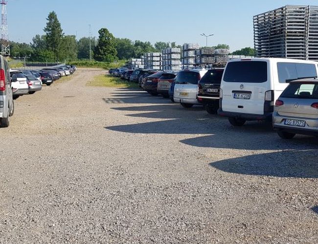 BaliceLot24 – Parking żwirowy pełen samochodów. Teren otwarty, z drzewami w tle. Po prawej stronie widoczne stosy palet.