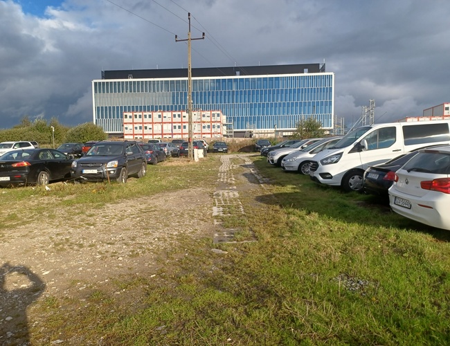 Maxi Parking - los coches están aparcados en un terreno de césped y grava. Al fondo se puede ver un edificio moderno con ventanas azules.