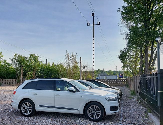 Parking MPark – varios coches aparcados en una superficie de grava. El área está cercada con una valla de metal. En el fondo se pueden ver árboles y cielo.