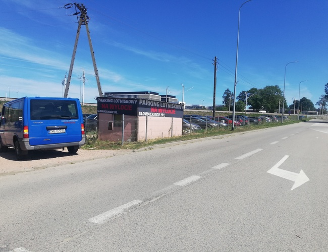 Parkovanie pri východe - Pred budovou stojí modré auto. Vedľa sa nachádza oplotené parkovisko. Na pozadí sú vidieť stromy.