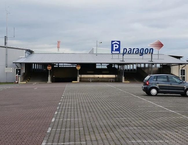 Widok na budynek z parkingiem przed wejściem. Na elewacji znajduje się oznakowanie parkingu, a przed budynkiem widoczne są miejsca postojowe i pojedyncze samochody.