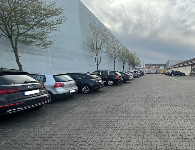 Parkovisko Mein-letisko-parkovisko-Frankfurt P2 – rad zaparkovaných áut. Vedľa stojí budova s stromami. Počasie je zamračené.