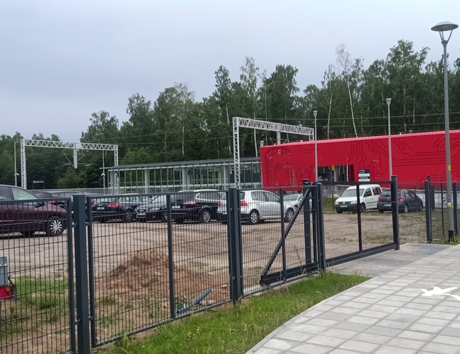 Inteligentné parkovanie – V pozadí je červená budova. Na parkovisku stoja autá. Pozemok je ohradený kovovým plotom.