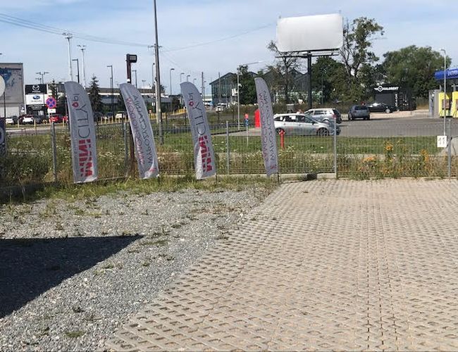 Auto Airport Samoobsługowy – Parking jest ogrodzony metalową siatką. Teren wyłożony jest betonowymi płytami i żwirem. Znajdują się tu flagi reklamowe.