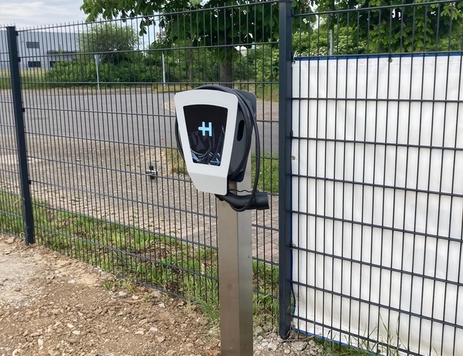 Parking P365 Valet – Teren ogrodzony z bramą. Jest ładowarka samochodowa. W tle widać drzewa.