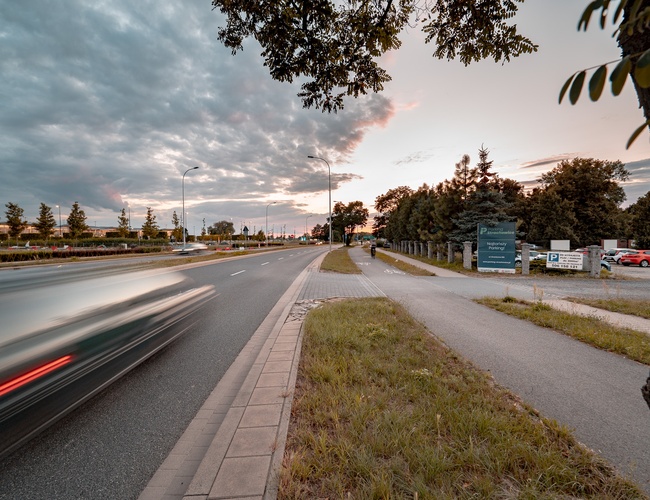 Parking Strachowice – Obok drogi znajduje się parking z drzewami. Ruchliwa ulica mija duże ogrodzenie przy znakach informacyjnych.