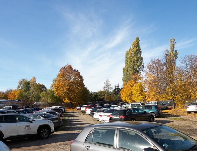 Parkuj Leć - Parking jest pełen samochodów, otoczony drzewami jesienią. Teren jest nieutwardzony i oświetlony.