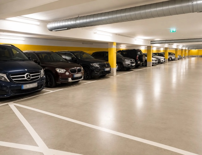 Podziemny parking z zaparkowanymi samochodami wzdłuż ściany, dobrze oświetlony przestronny korytarz.