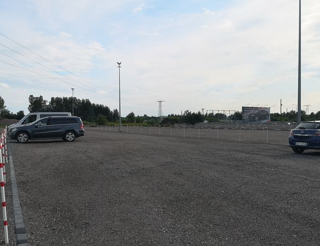 Parking A1 Rondo - Duży, otwarty plac z żwirową nawierzchnią. Kilka samochodów stoi zaparkowanych. W tle widoczne są drzewa i reklama.