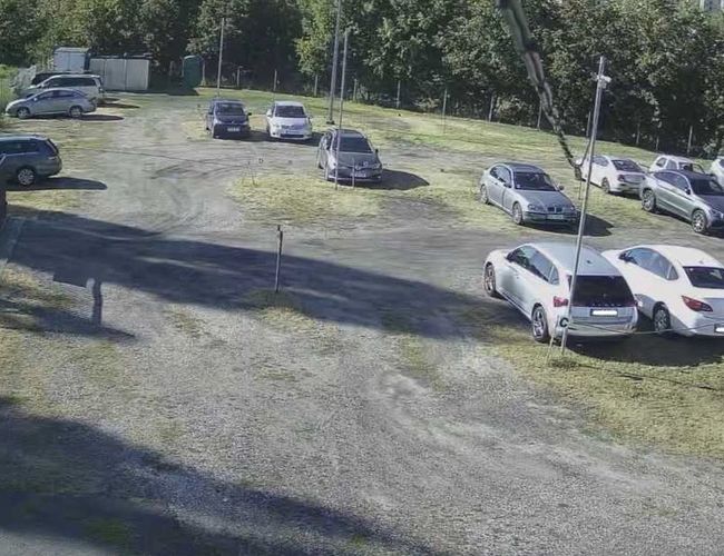 Estacionamiento de Salida BZG - El área no está cercada y es espaciosa. Se pueden ver coches estacionados. Al fondo crecen algunos árboles.