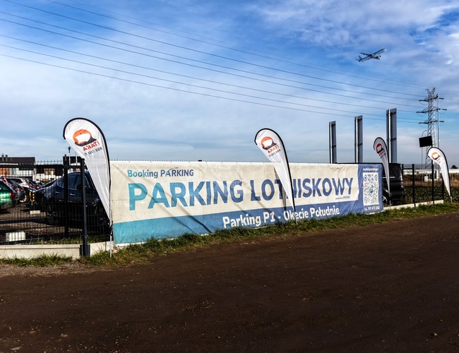 Parkovisko Okęcie juh - Oplotený pozemok s mnohými zaparkovanými autami. Na oplotení je veľký informačný banér.