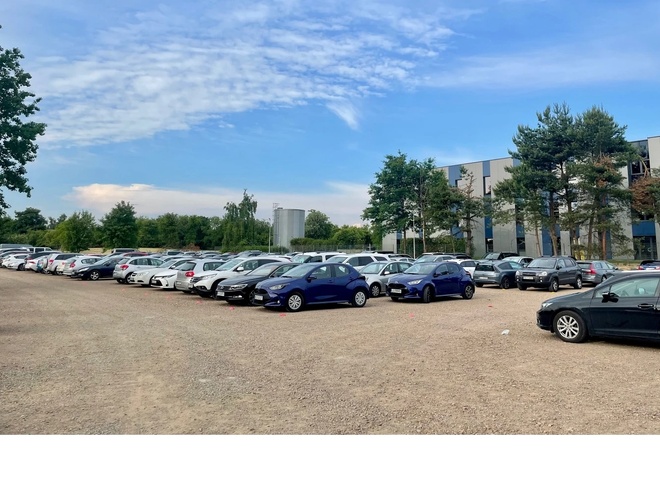 Sky Parking - Parking z wieloma samochodami na żwirowej nawierzchni. Otoczony jest drzewami i budynkiem z tyłu.