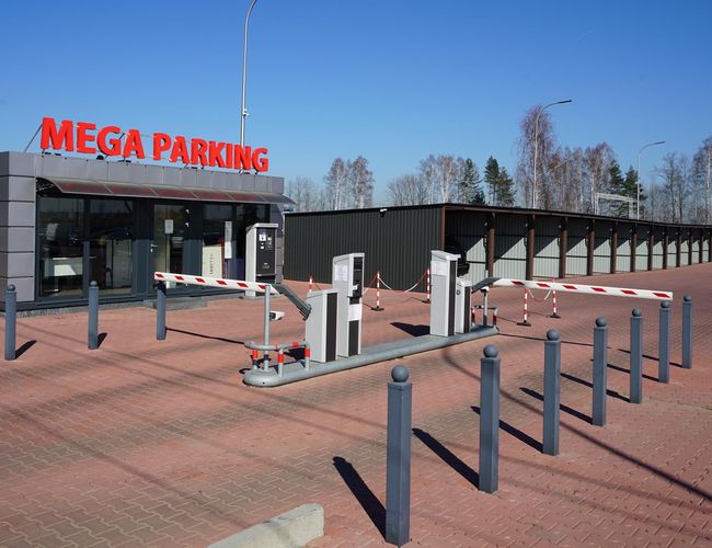 MEGA Parking - En la plaza hay un puesto con el logo del parking. Al lado están las barreras y la entrada al parking. Al fondo se ven los espacios de estacionamiento techados.