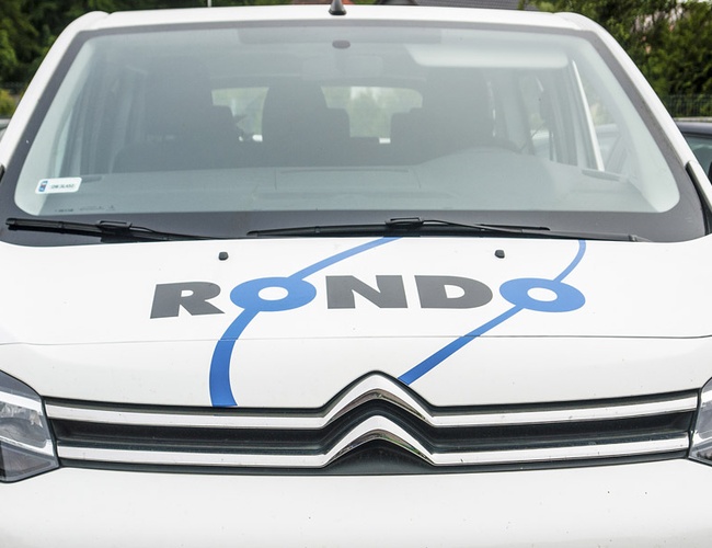 Parking Rondo – białe auto z logo Rondo na masce. Auto stoi na parkingu. W tle są drzewa i ogrodzenie.