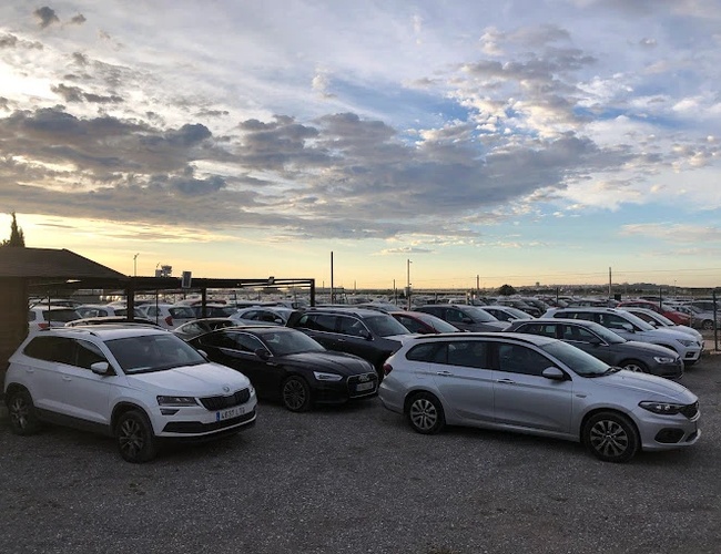 Sunline Parking Outdoor Valet – Viditeľné parkovisko s osobnými vozidlami. Pozemok je otvorený a kamenistý. Na obzore je obloha s mrakmi.