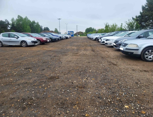 Primer Aparcamiento – Fila de coches aparcados sobre superficie de grava. A la izquierda y a la derecha hay coches. Al fondo se ve vegetación.