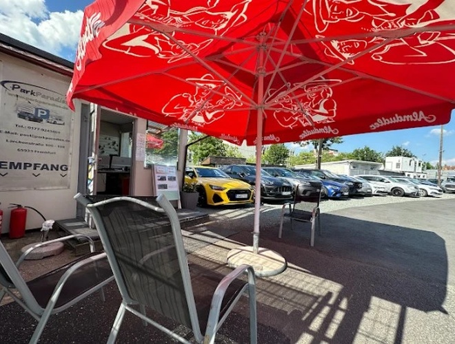 Parkhaus Parkparadies - Großer Parkplatz mit zahlreichen Autos. Roter Sonnenschirm und Stühle stehen neben der Rezeption.