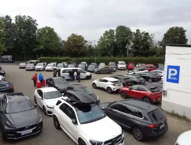 Parking Park&Fly Düsseldorf - Na parkingu stoi wiele aut. Ludzie poruszają się między nimi. Obok jest tablica z napisem "P 24h".