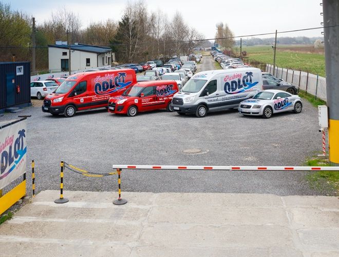 Parking Odlot - Na parkingu stoją zaparkowane samochody i dwa duże pojazdy firmowe. Teren jest ogrodzony i zamknięty szlabanem.