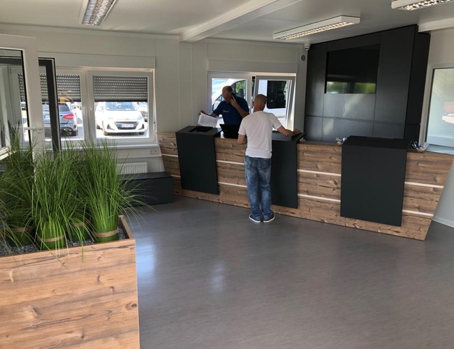 Tvoje miesto na parkovanie – Recepcia s dreveným pultom a rastlinami. Zákazník hovorí s pracovníkom. Na pozadí sú viditeľné okná a auto.