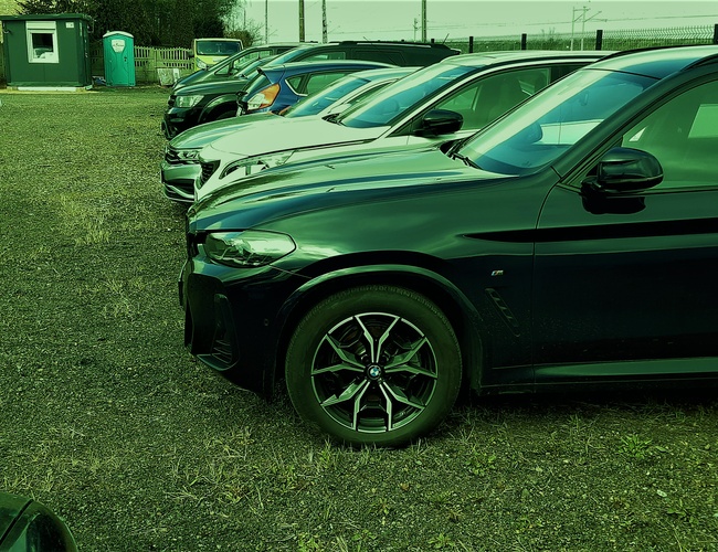 Gran Estacionamiento - Área con coches aparcados en la hierba. Al fondo hay una caseta y un baño portátil. El estacionamiento está cercado.