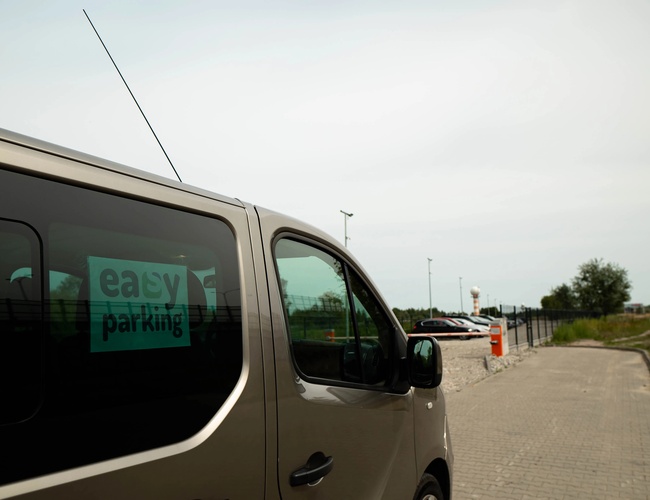 Easy Parking Express - Minivan s logom vstupuje na parkovisko. Vedľa je plot a pouličné svetlá. V pozadí je niekoľko zaparkovaných áut.