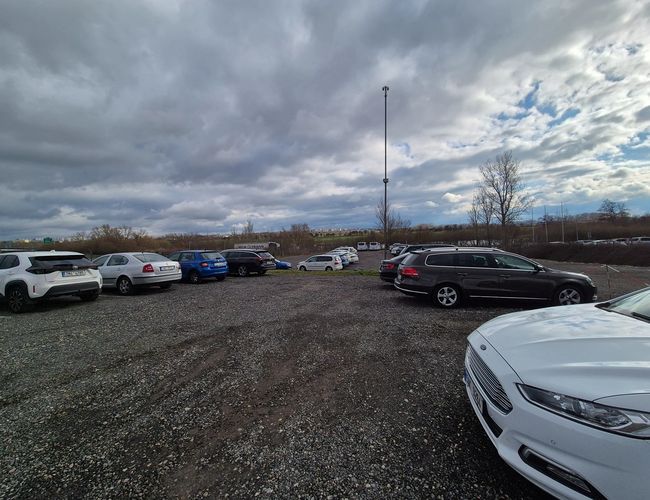 HostParking - Na żwirowym terenie stoi kilka aut osobowych. Parking jest otoczony drzewami. Na horyzoncie widnieje zachmurzone niebo.