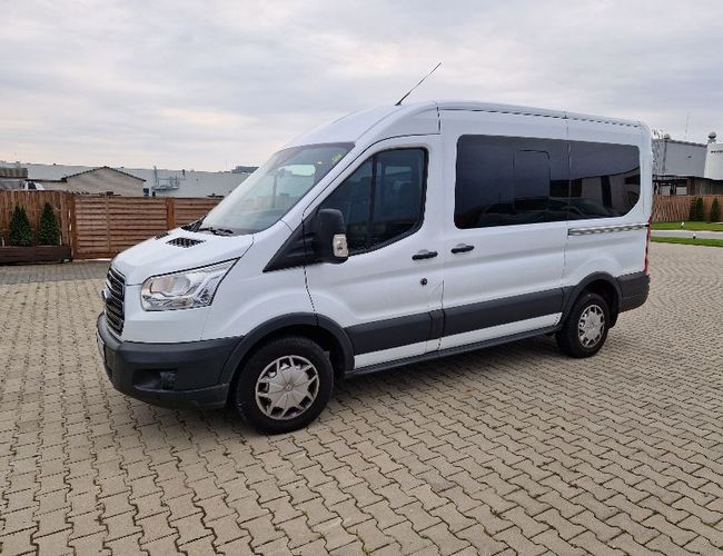 Parking Dominik – Biały bus stoi na brukowanym placu. W tle widać drewniane ogrodzenie i budynki.