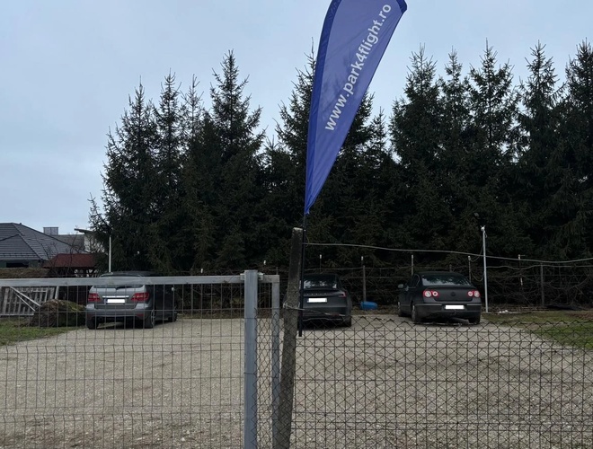Ogrodzony parking Park4flight Maramureș z widoczną niebieską flagą reklamową przy bramie wjazdowej oraz kilkoma zaparkowanymi samochodami na żwirowym terenie, w otoczeniu wysokich drzew.