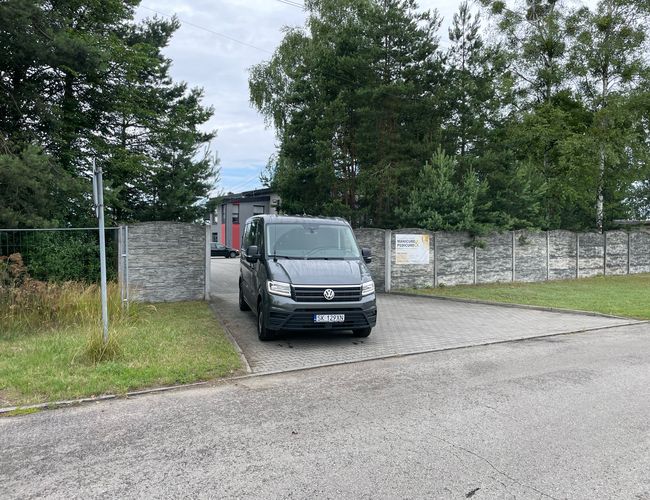 Parking Port – Wjazd z bramą i szarym autem. Otoczenie zieleni, trawa i drzewa.