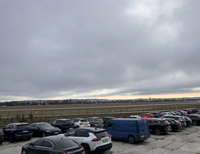 Parking MB Park – Zaparkowane są różne samochody osobowe i dostawcze. Teren jest ogrodzony i utwardzony płytami betonowymi. W tle widać pola.