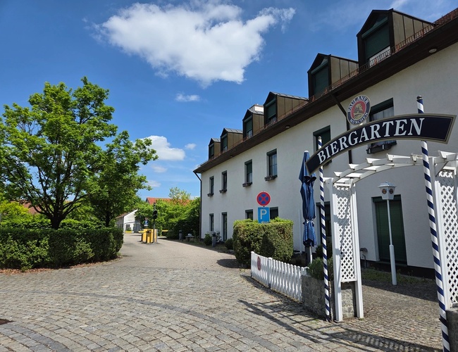 Atomis Hotel Munich Airport Parking - Edificios blancos con el letrero "Biergarten". Al lado se puede ver una acera y vegetación.