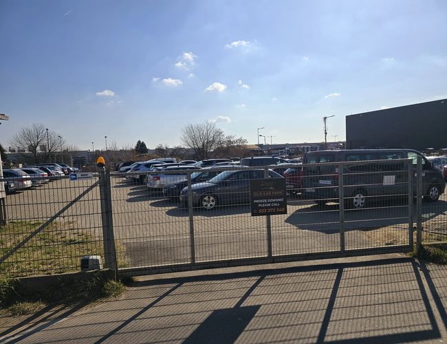 Parking Olo Car Park - ogrodzony, jasny plac z zaparkowanymi samochodami. Wejście zabezpieczone bramą. Panuje słoneczna pogoda.