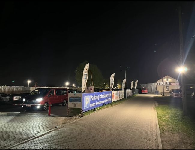 Parking Blisko Lotniska – widoczne duże flagi i tablice informacyjne. Kilka zaparkowanych samochodów na utwardzonym terenie. Sunny dzień, czyste niebo.
