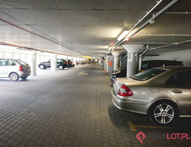 Oficjalny Parking Lotniska P2 – parking podziemny z wielu miejscami. Kilka zaparkowanych samochodów znajduje się przy betonowych filarach. Światło jest sztuczne, ściany są szare.